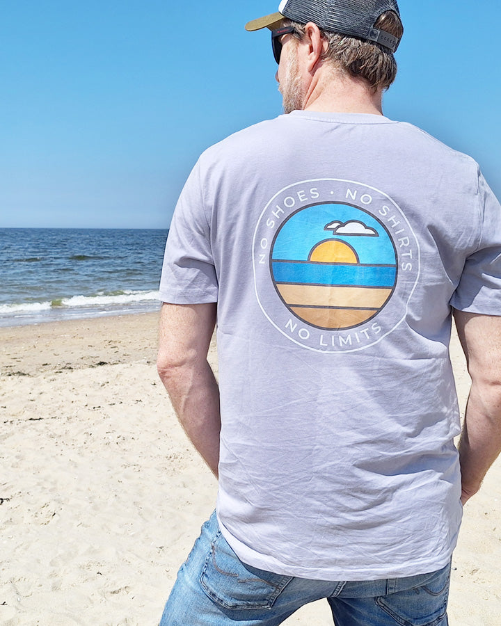 Beachvolleyball Bekleidung Streetwear Sportswear Oberteil Tshirt lavendel mit Markenlabel Großer Rückenprint Sonnenuntergang im Kreis No Shoes No Shirts No Limits Aufschrift Mann am Strand von hinten mit Shirt Sand Sonne Sommer